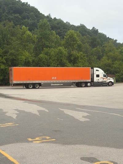 An orange semi-trailer truck is parked on an asphalt lot in front of a dense line of trees on a cloudy day. The truck is white on the cab and orange on the trailer. The trailer is long and rectangular, with reflective diamond shapes on the rear. The text "SchneiderJobs.com" is visible on the side of the trailer. The asphalt lot has yellow painted markings and some wet patches that cast shadows. The background is a heavily wooded hillside.