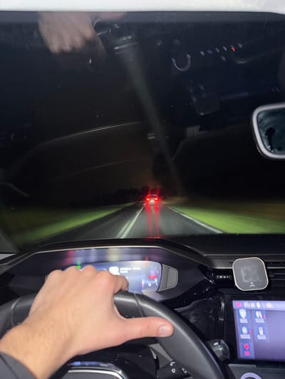 From the driver's perspective, a car travels on a road at night. The vehicle is directly behind another car, visible by its red taillights. The driver's hands are on the steering wheel, and the car's dashboard, including the digital display and rearview mirror, are visible. The road is dark, with faint green edges suggesting grassy or wooded areas on either side.