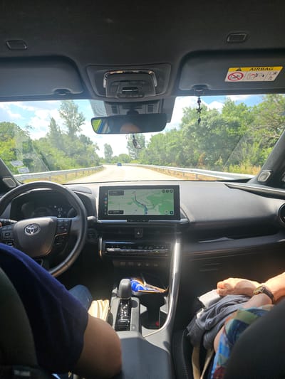 This media file depicts the interior of a car, seen from the perspective of a passenger or the driver. The scene is oriented vertically in the image. The car is in motion, as indicated by the view through the windshield showing a road curving through a landscape of trees and green foliage. The sky is visible, with some clouds, suggesting daytime.
Prominent objects include the dashboard, steering wheel with a Toyota logo, a central touchscreen display showing a navigation map, and various car controls. There is a sun visor with an "AIRBAG" warning label. A decorative item with stars and beads hangs from the visor. A person's arm and shoulder, wearing a dark blue top, are visible in the lower right portion of the frame. Another person's hands, wearing a watch, are visible in the upper right, appearing to hold or adjust some clothing.
Text visible in the image includes "AIRBAG", "0", "30 km", "3 hr 22 min", "VOL- VOL", "22.0", and "79". The navigation screen shows a map with a route and estimated travel time. The location context provided is Kesarevo, Bulgaria, suggesting the car is traveling in that region. The overall mood is neutral, capturing a moment during a car journey.