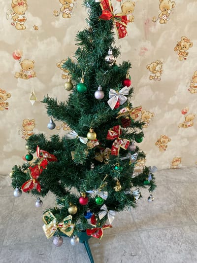 The image shows a close-up, top-down view of a decorated Christmas tree, likely an artificial one, with a light-colored wallpaper featuring cartoon teddy bears in the background. The tree is adorned with various ornaments, including red, green, gold, and silver baubles. There are also several decorative bows, primarily red with gold accents and some with a Christmas-themed pattern, and silver bows. A pine cone is visible among the branches.
The setting appears to be indoors, given the wallpaper and what looks like a tiled floor in the lower right corner of the image. The lighting suggests it could be daytime, but the exact time of day is not discernible. The overall impression is that of holiday decoration, likely for Christmas. No people are visible in the image. There is no visible text. The location context of Shostka, Ukraine, is provided but not directly evident from the visual cues in the image itself.