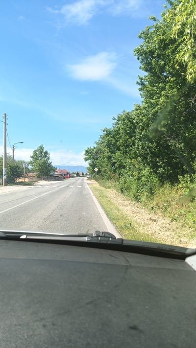 The image depicts a view from inside a vehicle traveling on a paved road. The location is Seltsi, Bulgaria. The car's dashboard and windshield obstruct the lower portion of the frame. The road is straight and appears to lead into the distance. On the left side of the road, there are houses and buildings, along with a utility pole. On the right side of the road, there is dense green vegetation. There is a diamond-shaped road sign on the right side of the road, closer to the vehicle. The sky is blue with some scattered clouds. Mountains can be seen in the far distance. There are no people visible in the image.
