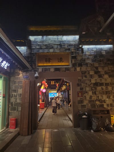 The image captures a nighttime scene in Wuhou District, China, likely a pedestrian walkway lined with traditional architecture. A prominent brick wall, possibly part of an ancient structure or a themed development, features a horizontal sign with Chinese characters. Below this sign, an arched entryway leads into a dimly lit area adorned with red lanterns and further architectural details. A CCTV camera is mounted on the wall near the entrance. On the left side, a storefront with illuminated signage is visible, and a red fire extinguisher is placed on the ground. The walkway is paved with stone slabs and shows reflections from overhead lighting. Several individuals are walking through the archway and along the path, their activities not clearly discernible due to the low light. Trash receptacles and overflowing plastic bags are situated on the right side of the walkway, adjacent to the brick wall. The overall atmosphere suggests a historic or cultural district open for evening strolls and commerce.