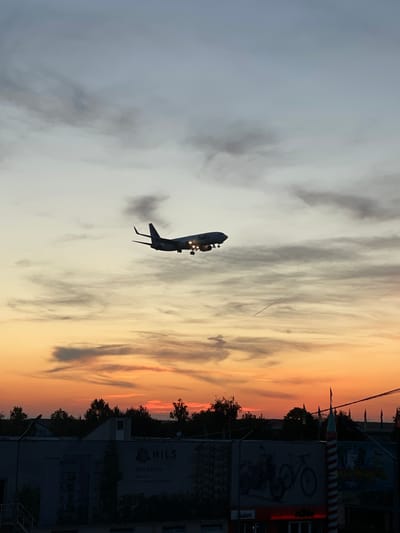An airplane is captured in flight against a vibrant sunset sky, with the image rotated 90 degrees clockwise. When correctly oriented, the aircraft is flying horizontally from left to right. The sky transitions from a soft grey-blue on the left to warm hues of orange, yellow, and pink on the right, indicating either sunrise or sunset. Scattered clouds are present, some reflecting the warm light.

The airplane itself appears as a dark silhouette, possibly a passenger jet, with its landing gear visibly extended and bright lights illuminated on its underside. A faint trail of exhaust or vapor is visible behind one of its engines. The low altitude suggests the plane is either on approach for landing or has recently taken off.

Below the horizon, the foreground features are largely in shadow, silhouetted against the bright sky. A line of trees runs along the horizon. To the right, various buildings and commercial signs are visible.

Visible text and details on the signs include:
*   A prominent sign displaying "HILS", along with a phone number "0310 052 174", a website "www.hils.ro", and an email address "e-mail: hils@hils-ro.eu".
*   Another sign shows the logo "KTM" and a graphic of a bicycle or motorcycle.
*   Partially visible text suggests "WATERPARK" and there is a cartoonish character, possibly a clown, on another advertisement.

The scene combines the natural beauty of a cloudy sunset with the man-made elements of air travel and urban development, capturing a fleeting moment near an airport. The vibrant sky contrasts with the dark, silhouetted subjects.
