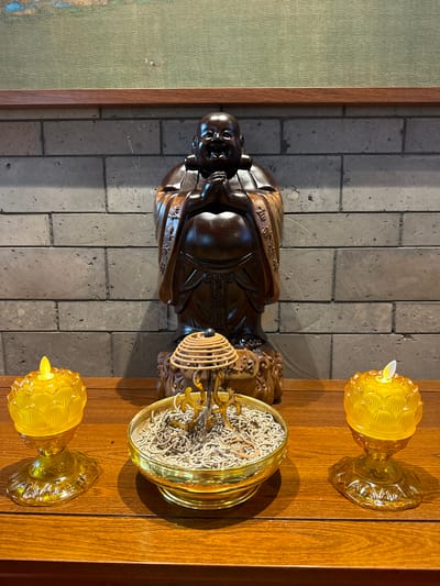 An upright-oriented indoor scene displays a collection of spiritual objects arranged on a polished wooden surface, likely a table or shelf, against a textured gray brick wall.

The central subject is a dark brown, intricately carved wooden statue of a smiling Buddhist monk or deity, possibly Hotei (Budai), depicted with a jovial expression and traditional robes. Chinese characters, including "福" (fú - good fortune/blessing), are faintly visible carved into the robes. The statue rests on a decorative base, also carved from wood.

To the right of the statue, on the wooden surface, are two yellow glass candle holders, each designed in a floral or lotus pattern, holding a small, white, battery-operated LED candle simulating a flame. Between these candle holders is a round, golden metal bowl or censer, filled with ash and numerous short, white remnants of burned incense sticks or coils. A spiral-shaped, light-colored wooden or rattan incense holder is placed within this censer.

The background features a wall composed of vertically stacked, rectangular gray bricks or stones, showing some wear and texture. To the far left, a framed piece of artwork or textile is partially visible, characterized by a muted green, textured surface and bordered by a simple wooden frame.

Natural light appears to enter from the right, casting soft shadows of the objects onto the wall and wooden surface, suggesting daytime. The arrangement suggests a setting dedicated to worship or meditation, such as a home altar or a temple.