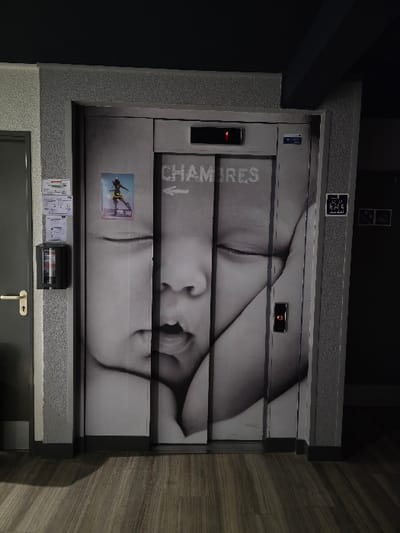 An elevator door displays a segmented black and white image of a woman's face. "CHAMBRES" is printed with an arrow pointing left. A small action sports advertisement and multiple informational labels are affixed below. The door frame is a speckled gray, with a dark gray door at the bottom. A horizontal door handle is present on the bottom door.