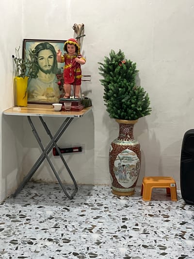 The image depicts an altar or shrine area situated indoors, likely in a residential setting in Koronadal City, Philippines. On a folding table, a framed portrait of Jesus is displayed, featuring his face and shoulders with a solemn expression. To the right of the portrait stands a small figurine of a child, possibly representing the Child Jesus, adorned in a red and gold garment and holding a cross. Beside these religious items, there's a yellow vase containing green foliage, a small white cup, and a shot glass with a clear liquid.
To the right of the table, a large, ornate vase with a predominantly reddish-brown hue is filled with a miniature artificial Christmas tree. The vase features intricate patterns and a painted scene depicting figures in traditional attire. In front of the vase, a small orange plastic stool is placed on a floor covered with terrazzo-style tiles, characterized by a mix of white, gray, and dark speckled fragments. A black speaker unit is partially visible on the far right of the frame. A red flashlight is attached to the underside of the folding table. The overall environment appears to be a corner of a room with plain white walls.