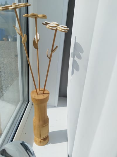 The image shows a wooden vase holding three decorative wooden flowers with yellow petals and light brown stems. The vase is positioned on a white windowsill. To the left of the vase, a window offers a view of an outdoor area with gravel and some structures. To the right, a white curtain is draped, casting shadows of the flowers onto its surface. The scene is illuminated by bright sunlight, creating distinct shadows. The overall impression is one of a simple, decorative arrangement placed near a window. There is no indication of the specific location within Bezlesnyy, Russia.