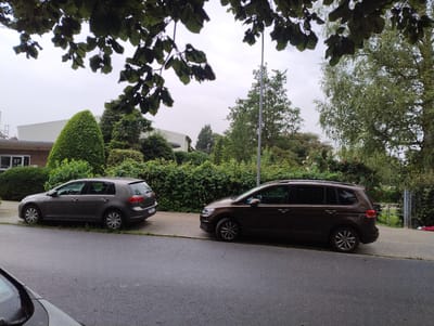 The image depicts a street scene in Uetersen, Germany, likely in a residential or semi-residential area. Two cars are parked on the right side of the asphalt road, facing towards the right of the frame. The car closer to the foreground is a dark brown multi-purpose vehicle, possibly a Volkswagen Touran, with its rear end visible. It is parked on a paved sidewalk next to a dense green hedge. To its left, further down the street, is a dark grey hatchback, likely a Volkswagen Golf, also parked on the sidewalk. Behind the cars and the hedge, several trees and bushes are visible, contributing to a verdant environment. A building, possibly a house or a small commercial establishment with the number "27" visible on it, can be seen behind the grey car, partially obscured by foliage. The sky is overcast, suggesting a cloudy day. The foreground of the image shows a portion of another car's windshield and hood, indicating the photo was taken from another vehicle. The overall impression is a quiet, everyday scene in a German town.