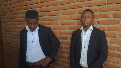 Two young men stand in front of a brick wall. Both wear white shirts, black blazers, and dark pants. The man on the left looks down with his left hand on his hip, a brown belt visible. The man on the right looks directly at the viewer. The background brick wall is a mix of orange and brown hues.