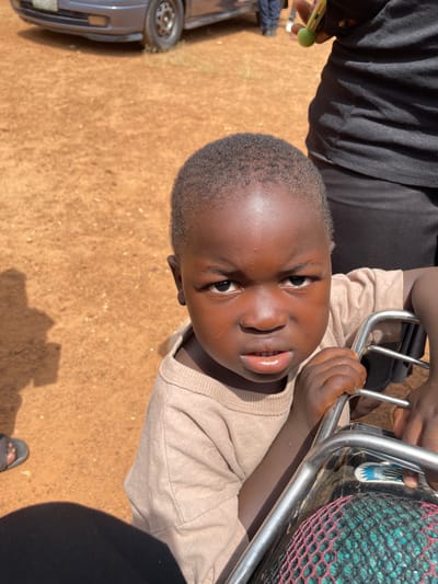 In Kassa, Nigeria, a close-up shot focuses on a young child with dark skin and short, curly hair, wearing a light brown t-shirt. The child has a serious expression, with furrowed brows and slightly parted lips revealing teeth. The child's hands are gripping a metal bar of what appears to be a motorcycle or similar vehicle. Behind the child, an adult wearing a black t-shirt and pants is partially visible, holding a green object. The background consists of reddish-brown dirt and dry grass. In the upper left corner, the front of a gray car is visible. The lighting suggests it is daytime, casting shadows on the ground.