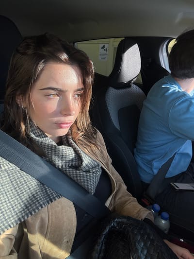The image depicts the interior of a modern vehicle during daylight hours in Mafra, Portugal. A young adult female is seated in the front passenger seat, positioned on the left side of the frame. She has brown, wavy hair and light-colored eyes. Direct sunlight illuminates the left side of her face from the vehicle's right side. She wears a light brown or tan suede-like jacket over a dark top, with a grey and white houndstooth pattern scarf around her neck. A dark seatbelt extends across her chest. A black woven-texture bag is on her lap, next to two small plastic water bottles with blue caps. A silver hoop earring is visible on her left ear. To her right, partially visible from the back, is a male individual occupying the driver's seat, wearing a light blue short-sleeved shirt. A tablet or smartphone with an illuminated screen rests near his lap. Through the passenger window, a light-colored exterior building wall is visible, featuring a white rectangular sign with a black surveillance camera icon and Portuguese text.