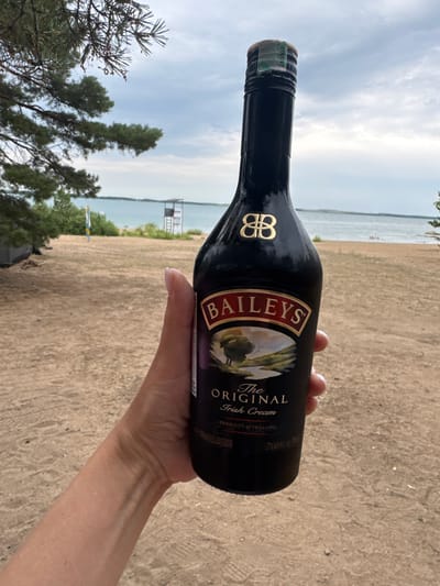 A hand holds a bottle of Baileys Irish Cream in front of a beach with a body of water in the background. The sky is overcast. In the distance, there is a lifeguard tower and a few people wading in the water. The ground is sandy and there are some trees to the left of the frame. The scene appears to be set in the city of Aiyrtau, Kazakhstan.