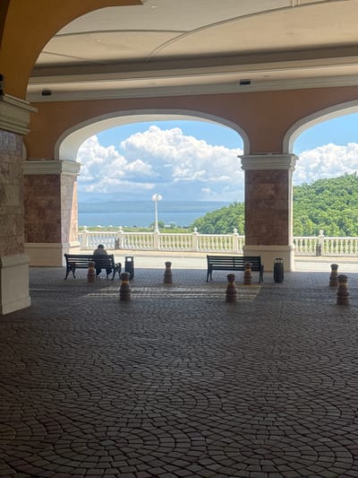 The image depicts a view through an arched walkway towards a body of water and a lush green hillside. In the foreground, a person is seated on a bench, facing away from the camera, looking out at the scenery. The ground is paved with interlocking stones arranged in a circular pattern. Several bollards are also visible on the paving. Beyond the walkway, there is a white railing overlooking the water, with trees and hills in the distance. The sky is filled with dramatic white clouds. The scene appears to be outdoors in a well-maintained area, possibly a public space or resort. There is no indication of the specific location within Shkotovo, Russia.