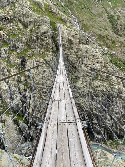 The image depicts the Trift Bridge, a pedestrian suspension bridge near Innertkirchen, Switzerland, observed from a perspective on its wooden plank walkway, looking longitudinally towards the opposite side of the gorge. The bridge features a walkway composed of light-colored, weathered wooden planks secured by metal bolts, flanked by robust steel suspension cables and a network of diagonal and vertical steel safety wires forming a cage-like structure. Metal clamps and brackets secure the cable system to the walkway.

Two human figures are discernible on the bridge approximately one-third of the way across, walking in the direction of the bridge's span. Their individual features and attire are indistinct due to distance.

The bridge spans a deep, V-shaped gorge characterized by steep, rugged rock faces on both sides. The rocks are predominantly grey and brown, with patches of green moss and sparse herbaceous vegetation adhering to crevices and ledges. On the right-hand side of the gorge, a visible stream of white water cascades down the rocky slope, feeding into a milky turquoise river that flows at the bottom of the chasm beneath the bridge. The scene is illuminated by diffuse natural light, suggesting an overcast sky. The overall environment is mountainous and alpine.