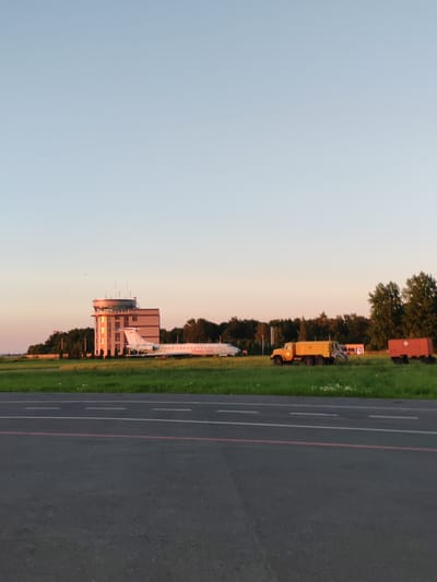 The image depicts an airport tarmac in Khrabrovo, Russia, during what appears to be sunset or sunrise due to the warm, golden light reflecting off the control tower and the clear, pale blue sky. In the mid-ground, a white passenger jet with visible lettering on its side is parked on the grass beside a multi-story control tower. The tower is light-colored with a dark roof and numerous windows, some of which are illuminated. To the right of the airplane, a bright yellow utility truck is parked on the grass, and further to the right, a red trailer is also visible. The foreground shows a portion of the airport tarmac with white runway markings and a thin red line. Trees line the background, creating a natural border behind the airport infrastructure. No people are visible in the scene, and the overall impression is one of a quiet, operational airport at the fringes of daylight.