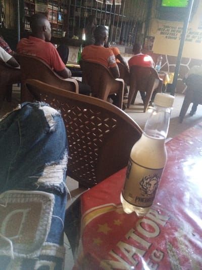 The image depicts an indoor scene within a social establishment, likely a bar or restaurant, located in Majengo ward, Kenya. In the foreground, the upper leg of an individual wearing distressed blue jeans with pronounced rips and white threads is visible on the left. A light-colored patch with an embossed, partially visible letter is present on the jeans. To the right, a clear plastic bottle with a white screw cap, containing a pale yellow liquid and showing condensation, rests on a table. The bottle features a gold label with a tiger's head graphic and the word "PREDATOR." The table is covered by a red plastic sheet emblazoned with white stars and text, including "NATOR" and "LEG," consistent with "SENATOR LAGER" branding. A brown plastic chair backrest with a perforated diamond pattern occupies the central mid-foreground.

In the midground, several individuals, appearing predominantly male, are seated in similar brown plastic chairs, facing away from the camera. One person wears a plain orange t-shirt, another an orange shirt with white sleeve stripes, and a third a red t-shirt. Small tables are interspersed, holding objects such as a green bottle and a clear plastic water bottle.

The background features a bar area behind a dark metal grate, with multiple shelves stocked with bottles. A yellow poster with black text in an unidentified script is attached to the right wall, which has a patterned stone or tiled lower section. Above the poster, a television screen displays a green-dominated image, suggestive of a sports broadcast. The environment is illuminated, with some reflective glare on the table surface.