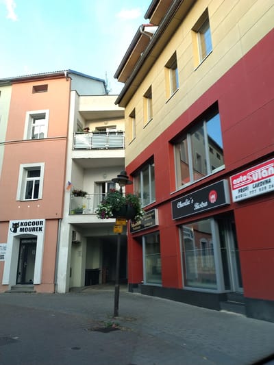 The image shows a street scene in Strakonice, Czechia. The view is from a street level, looking up at several buildings. On the left is a light pink building with a white-framed window and a shopfront with a sign featuring a black cat logo and the text "KOCOUR MOUREK". To the right of this is a white building with a balcony. Further to the right, a building is painted in a striking red and yellow combination. This building features several windows and a sign for "Charlie's Bistro". Another sign on the red building reads "auto Sulan". The street is paved with grey interlocking bricks. A lamppost with hanging plants is visible in the foreground. The sky above is blue with some scattered clouds. The scene appears to be during daylight hours, possibly late afternoon or early evening, given the lighting.