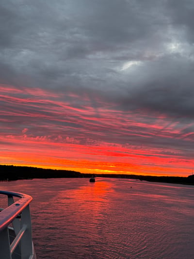 A wide-angle, horizontal, exterior shot captures a vibrant sunset over the Svir River in Voznesenye, Russia. The sky is filled with streaks of fiery orange and deep red clouds, contrasted by darker, grey clouds above. The river, reflecting the intense colors of the sky, appears as a broad expanse of rippling water. In the distance, a dark, tree-lined shore runs along the horizon, with a few small structures and a tall, slender tower visible on the right side of the frame. A few boats are also present on the water, including a larger vessel in the distance. In the foreground, a white railing with a blue top edge suggests the viewpoint is from a ship or ferry. The overall mood is serene and dramatic, dominated by the spectacle of the setting sun.