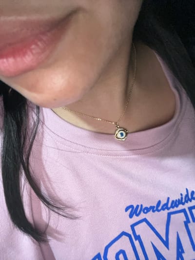 The image shows a close-up of a person's neck and chest area, focusing on a gold necklace with a blue and white evil eye pendant. The person is wearing a light pink t-shirt with blue text that says "Worldwide" and part of another word. The background is dark. The image does not contain any information or context related to Conuco Viejo, Venezuela.