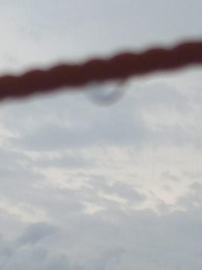 The media file presents an abstract, blurred composition dominated by a dark, vertical, elongated object in the foreground, likely a rope, cable, or similar structure, extending upwards and downwards out of the frame. This object is significantly out of focus, appearing soft and indistinct.

The background consists of a cloudy sky, rendered in varying shades of white and light grey-blue, suggesting a daytime scene with overcast weather. To the right of the dark foreground object, approximately in the upper-middle section of the sky, a very faint, blurred, and somewhat circular or semi-circular shape is discernible.

No people, discernible activities, or specific events are taking place. The image is static, focusing on environmental elements. There are no visible texts or specific location cues within the image itself to identify a particular place, though the provided context indicates Puloblang, Indonesia. The overall impression is one of soft focus, possibly an accidental shot or an artistic rendering emphasizing texture and light.