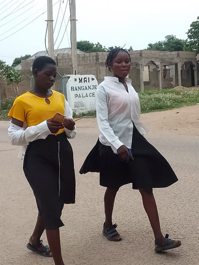 Two young women are walking along a paved road in Biliri, Nigeria. One woman is wearing a yellow top with a white over-shirt and a black skirt with a zipper down the front. She is looking down at a phone in her hand. The other woman is wearing a white blouse and black skirt. Both women are wearing dark sandals. In the background there is an unfinished building and a sign that reads "Mai Banganje Palace". Utility poles with wires are also visible.