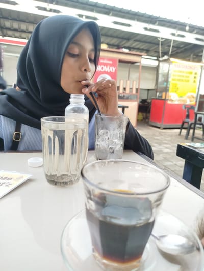 A young woman wearing a black hijab and a blue and white striped shirt is seated at a white table. She is drinking from a black straw. In front of her are two glasses, one filled with water and the other with ice. A clear plastic bottle of water is also on the table, along with a small white cap. To her right, a cup of dark liquid, likely coffee, sits on a saucer with a spoon. In the background, there are food stalls with colorful signage, suggesting an outdoor food court or market. The scene appears to be a casual meal or break.