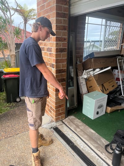 The image depicts a young adult male standing outside a brick garage entrance in Wombarra, Australia, on an overcast day. The subject, positioned with his right side partially facing the viewer, wears a dark blue t-shirt, tan cargo shorts, a black baseball cap, and tan work boots. His right arm is extended downwards, with his index finger pointing towards a metal drainage grate embedded in the concrete ground directly in front of the garage threshold. He holds a small, orange, spherical object in his right hand. A green utility tool is clipped to his right shorts pocket.

The garage entrance, partially open with a rolled-up metal door, reveals an interior with a green floor covering, possibly artificial turf. Inside, numerous cardboard boxes of varying sizes are stacked, along with a tall, silver-framed mirror leaning against a wall. Visible box labels include "MOMCOZY Auto Bottle Washer" and "Moka." A window with security bars is visible at the back of the garage, showing an overcast sky and residential rooftops.

To the left of the garage entrance, two wheelie bins are present: one dark grey with a yellow lid, and another dark grey with a red lid. Behind these, dense foliage, including a tree fern, is visible. On the green floor inside the garage, near the drainage grate, a black bag and a black water bottle are situated. The ground outside the garage is a pebbled concrete surface.