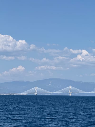 This media file depicts a panoramic view of a suspension bridge spanning across a body of water, with a mountainous landscape in the background. The scene is split horizontally, with the sky and clouds occupying the upper portion and the bridge, water, and land filling the lower half.
The sky is a clear, bright blue, punctuated by scattered white, puffy clouds. The sea is a deep, rich blue with a textured surface, indicating gentle waves or ripples. The bridge, a prominent object, is a modern suspension bridge with two tall, white pylons that support numerous cables forming a V-shape. The bridge extends across the frame, connecting the land on either side.
In the distance, hazy blue mountains rise behind the bridge. A distant shoreline with some buildings can be seen along the base of the mountains. The overall atmosphere is serene and majestic, suggesting a calm and pleasant day. The lighting indicates it is daytime, likely mid-morning or afternoon, with the sun illuminating the scene.
The location is identifiable as Patras, Greece, known for its impressive bridges connecting the Peloponnese peninsula to the mainland. The image captures the grandeur of the engineering feat against the natural beauty of the Greek landscape. No people are visible in the image, and there is no visible text.