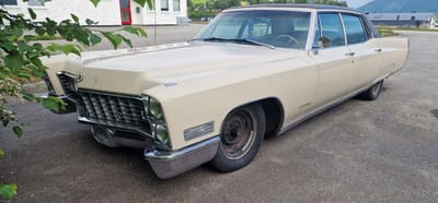 The image shows a vintage cream-colored Cadillac parked on an asphalt surface in Ørsta, Norway. The car is a four-door sedan with a black vinyl roof and chrome accents. The front of the car features a prominent grille, rectangular headlights, and a chrome bumper. The car's tires are black and appear to be older models.

In the background, a white building with a red door is visible, along with some trees and vegetation. Beyond that, there are houses and hills, suggesting a semi-rural or suburban setting. The sky is partly cloudy. The image seems to be taken from a low angle, looking up at the car. The overall impression is that of a classic car parked in a quiet Norwegian town.