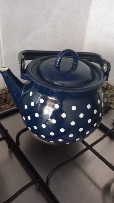 A close-up, eye-level shot shows a dark blue, polka-dotted enamel teapot resting on a black metal stovetop grate in a kitchen. The teapot has a white spout and a black handle. The stovetop is a light gray metallic color, and the background appears to be a tiled wall with a light, mottled pattern. The lighting suggests an indoor setting, possibly in a home in Oliveira do Bairro, Portugal. There are no people present in the image, and the teapot appears to be stationary.