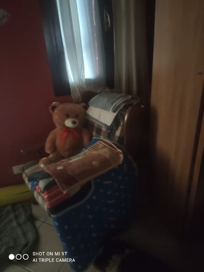 A fuzzy, low-light indoor scene depicts a teddy bear sitting on a chair piled with blankets. The teddy bear is brown with a red bow tie. The blankets appear to be stacked in a haphazard manner. A dimly lit room with red walls is visible in the background, along with a window covered by white curtains. The overall impression is of a cluttered, personal space. The location is specified as Bagnoli di Sopra, Italy.