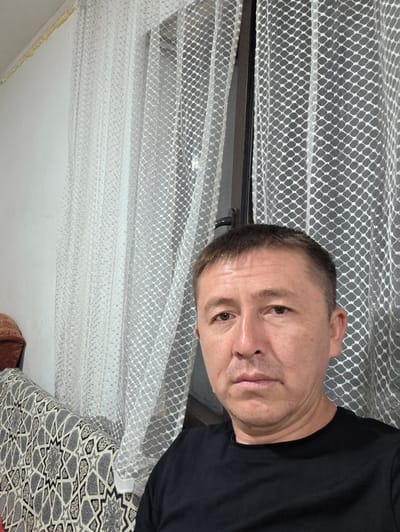 The image shows a close-up, selfie-style portrait of a middle-aged man with short, dark hair and brown eyes. He is wearing a black t-shirt and looking directly at the camera with a neutral expression. The background consists of a white wall on the left, a window with a white, patterned sheer curtain covering it, and a patterned sofa with a geometric design in shades of gray and black on the left side of the frame. The man appears to be indoors, possibly in a living room or common area. The lighting suggests an indoor setting with natural light coming from the window. There is no specific indication of the city of Kochkor, Kyrgyzstan, within the image itself.
