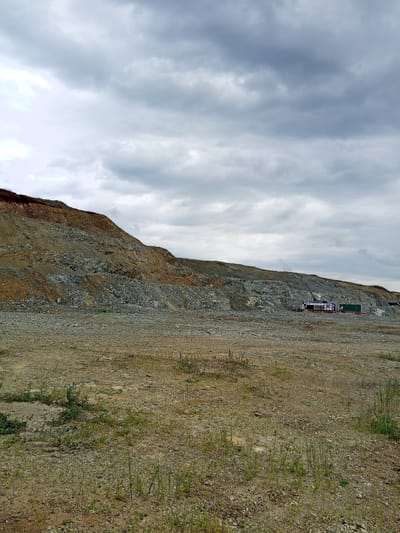 The image depicts an outdoor scene at what appears to be a construction or excavation site in Raygorodok, Kazakhstan. The foreground is a flat, barren expanse of gravel and dry earth, with sparse tufts of green grass growing sporadically. In the midground, a large, tiered rock face dominates the landscape, characterized by layers of reddish-brown earth and grey rock. The excavation cuts into the hillside, revealing distinct geological strata.
Further back, towards the right side of the image, there are several industrial structures. These include what appear to be prefabricated modular buildings or shipping containers, some white with dark roofs, and a large green container. A crane arm extends upwards from behind these structures, suggesting ongoing construction or operational activity. The overall environment is exposed and rugged, with evidence of significant earthmoving.
The sky above is overcast with heavy, grey clouds, contributing to a subdued and potentially humid atmosphere. There are no visible people in the frame. The scene is static, captured from a ground-level perspective, offering a wide view of the raw, uncultivated terrain and industrial elements within it.