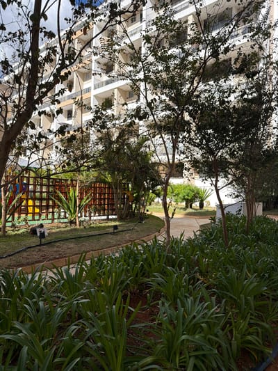 The scene is an outdoor view in what appears to be a residential area, likely in Brasília, Brazil given the architectural style. A tall, multi-story building with multiple balconies is visible through the branches of trees on the left side of the image. In the foreground, there is a well-maintained garden area with a variety of green plants. There is a wooden lattice fence with greenery and perhaps playground equipment visible behind it. A paved walkway extends into the distance, leading toward another building partially visible in the background. The light suggests a sunny day.