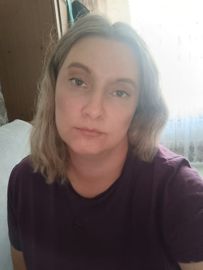 A woman with light blonde hair and fair skin is centered in the frame. She wears a purple T-shirt. Her expression is neutral. Behind her is a wooden cabinet to the left and a window with white patterned curtains to the right. The location is within a domestic environment. Closed administrative–territorial formation "Mezhgorye", Russia cannot be determined from the provided imagery.