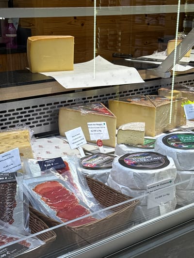 The image displays a brightly lit refrigerated display case, likely found in a deli, cheese shop, or a gourmet food store. The scene is filled with a variety of cheeses and some cured meats, neatly arranged for sale. The background behind the products appears to be a warm, wooden paneling, suggesting a rustic or traditional shop interior.
On the far left, a large, rectangular block of light yellow cheese, possibly unwrapped, stands upright on a display surface. To its right, a white sheet of paper with a subtle pattern is laid out, partially visible.
The central and right sections of the display case are packed with various products. Several large blocks of cheese, some wrapped in clear plastic, are visible. One prominent block, also wrapped in clear plastic, features a white label with red text, identifying it as "Meule des Bauges" and priced at "20,10€ Le Kg." Next to it, a wedge of cheese with a natural, greyish rind is displayed. Below this, another large block of cheese is visible.
Further to the right, several round cheeses are wrapped in white paper, each bearing a distinct label. One clearly states "TOMME CHAMARD," while another is labeled "Tomme du Châtelar" with a price of "14,40€ Le Kg." A third round cheese has a label that partially reads "TOMHE PELAA." These round cheeses are arranged on shelves, some resting in wicker baskets.
In the lower right portion of the display, vacuum-sealed packages of sliced cured meats are visible. One package of sliced ham is labeled "JAMBON" with a price of "8.50€." Below this, another package contains what appears to be sliced salami or a similar dark, cured sausage. Also, a block of cheese with visible holes, characteristic of Emmental, is labeled "EMMENTAL" and priced at "15,20€ Le Kg." Another label in this area reads "90 €."
The overall impression is one of a well-stocked and inviting food display, showcasing gourmet products, primarily cheeses, with prices listed in Euros, indicating a European location. No human activity is depicted in the static image.