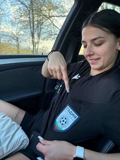 A woman is in a vehicle. She is wearing a black referee jersey with a patch that says "СЪДИЯ REFEREE" and includes the Bulgarian flag. She is pointing at the patch with her right index finger. She is wearing a white watch on her right wrist. Part of a tattoo is visible on her thigh. The view out of the vehicle window is blurry, but shows trees and the sky. The location is not apparent from the image. There is no information to suggest this photo was taken in Gorna Rositsa, Bulgaria.