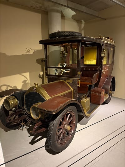 This image captures a vintage automobile, likely an early 20th-century model, displayed indoors, possibly within a museum or exhibition space in Wassenaar, Netherlands. The car is seen from a slightly elevated side angle, facing towards the right.
The vehicle is characterized by its dark brown bodywork and prominent brass accents, including headlights, trim, and interior fixtures. It features large wooden spoke wheels with dark pneumatic tires, one of which appears to be a spare mounted on the side. The front of the car shows a distinctive grille with a small, circular manufacturer's emblem containing stylized letters, possibly "L-F". A brass-colored radiator cap is visible above the grille.
The car's design suggests a coach-built body, possibly a landaulet or brougham, with a closed rear passenger compartment featuring glass windows and a more exposed front driver's area where a wooden steering wheel is visible. The interior of the passenger compartment shows hints of plush, possibly yellow, fabric or upholstery, and what might be a map or decorative lining through the windows. A small wooden box and a brass-colored container are secured on the running board or fender. A detailed, clear glass lantern or lamp is attached to the side of the cabin.
The setting consists of plain, light-colored walls—a beige wall on the left and a white, tiled wall on the right. Dark, thin lines, possibly ropes or barriers, stretch diagonally across the white wall. Prominent shadows cast by the car and other structural elements suggest strong artificial lighting. A white pipe or cylindrical fixture is visible along the left wall. No people are present, and the vehicle is stationary, serving as a static exhibit. No discernible text beyond the car's emblem is clearly legible.