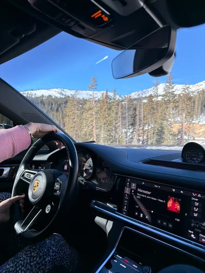 The image captures the interior of a car, focusing on the dashboard and steering wheel. A person's hands are visible on the wheel, suggesting they are driving. The car's infotainment screen displays the song "Maneater" by Daryl Hall & John Oates. Outside the windshield, a snowy mountain landscape with trees is visible under a clear blue sky. The dashboard features various controls and displays, indicative of a modern vehicle.