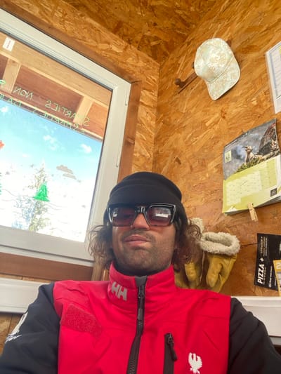 An adult male is captured in a close-up, frontal portrait indoors. He wears a black knit beanie, dark-framed sunglasses with a reddish tint, and a red jacket with black sleeves, bearing the Helly Hansen "HH" logo and a white phoenix-like emblem. His dark, curly hair is visible beneath the beanie. The interior walls and ceiling are constructed from oriented strand board (OSB). To the left, a white-framed window shows an outdoor scene featuring a clear blue sky, snow-covered ground, and evergreen trees. The window glass is adorned with reversed text such as "Smarties" and "non," along with green tree and brown cloud decals. To the right, a light-colored, patterned baseball cap hangs on a wooden peg. Below the cap, a wall calendar depicting an ibex on a mountain is visible, with French text. A pair of tan leather gloves with cream-colored fleece cuffs hang below the calendar, next to a partially visible black pizza menu attached with a wooden clothespin. The setting is consistent with a casual establishment, such as a cafe or snack bar, in the mountainous region of La Salle-les-Alpes, France.