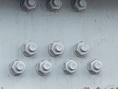 The image shows a close-up of a metallic surface with several hexagonal bolts. The bolts appear to be made of metal and are painted or coated in a light gray color, with some areas showing signs of wear and rust, particularly along the bottom edge of the frame where there is visible orange-brown corrosion.
The bolts are arranged in a grid-like pattern. The top row has three bolts, spaced relatively far apart. Below this, there are two more rows. The second row contains four bolts, and the third row contains four bolts. All bolts in the lower two rows are more closely clustered together than those in the top row. Each bolt consists of a hexagonal head with a washer-like feature beneath it, and likely a nut on the other side, though only the heads are clearly visible. The metallic surface itself is also painted gray and shows some minor surface imperfections and speckling.
There is no activity or event taking place; the image is a still shot of a static object. The lighting suggests it might be daytime, but there are no strong shadows to indicate a specific time of day. Weather conditions are not apparent. The metallic surface and the type of fasteners suggest it could be part of a structure like a bridge, building, or machinery. Given the provided location context of Parit Baru, Indonesia, this could be a component of infrastructure in that region. No text is visible in the image.
