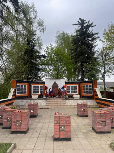 A monument is in an outdoor space in Хлябово, Russia. It consists of a white wall with a drawing of a soldier in an overcoat and a monument with two sets of six white rectangles on a black background. There are red brick structures with a single brick missing on top that are lined up in rows on the grey concrete. Wreaths and flower arrangements are placed on the monument and on the brick structures. Tall trees are around the monuments and buildings are on the background. The sky is grey.