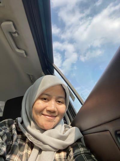 A woman wearing a light gray hijab and a plaid shirt is looking at the camera. She is smiling and her face is illuminated by sunlight. She appears to be sitting in a car, with part of the car's interior visible, including a handle on the ceiling and a brown leather-textured panel. The sky, with scattered clouds, is visible through the car's windows. The scene is outdoors on a bright day.