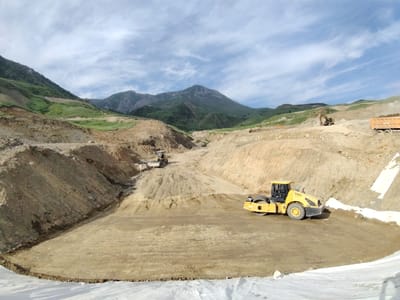 This is a landscape photograph taken at a construction site in Farab, Tajikistan. The scene depicts a large-scale earthmoving operation with heavy machinery at work. In the foreground, a yellow steamroller is compacting a dirt surface. In the middle ground, a yellow excavator is seen operating on a dirt slope, and further back, another yellow excavator is visible. A large orange dump truck with a load of material is parked on a higher level of the site, to the right of the frame.
The setting is a hilly, mountainous area, with the terrain heavily excavated and shaped by construction activities. The surrounding landscape features rugged, bare earth and slopes, interspersed with patches of green vegetation on the distant hillsides. The sky is blue with scattered white clouds, suggesting a clear day, likely during daylight hours. The overall impression is one of an active construction project in a remote, natural environment. There is no visible text in the image.