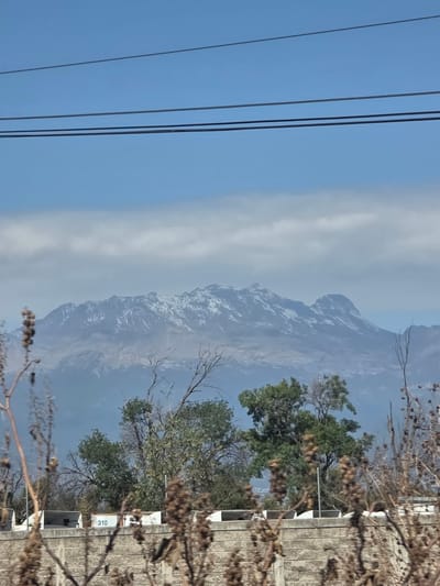 The image shows a mountainous landscape partially obscured by haze. The mountain range has snow-capped peaks. In the foreground, there are trees with green foliage and bare branches. Power lines stretch across the upper left portion of the frame. A building with the number "310" visible on its side is located on the right of the frame, partially obscured by branches. The location is Santa Ana Xalmimilulco, Mexico.