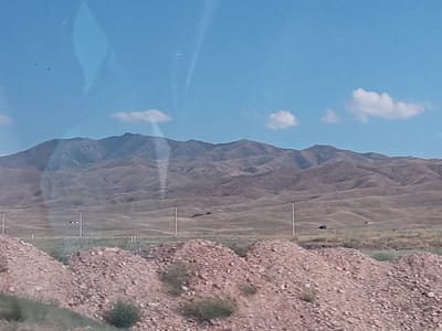 The image depicts a landscape scene with rolling hills in the background under a blue sky with scattered white clouds. In the foreground, there are piles of reddish-brown gravel or soil. In the middle ground, there are several utility poles with wires, and one pole has a rectangular black and white sign attached to it. The landscape appears arid, with dry vegetation visible between the gravel piles and on the hillsides. There are no people visible in the image. The scene is likely a rural or semi-rural area. The prompt specifies the location as the city of Bereke, Kazakhstan, but this specific detail cannot be visually confirmed from the provided image alone.