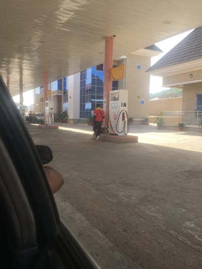The image depicts a gas station in Jiru, Nigeria, taken from inside a car. A person in a red shirt is filling a vehicle with gas at a pump. The station has modern architecture, with a mirrored building and a covered area for the pumps. The ground is concrete. The image is oriented in portrait mode.