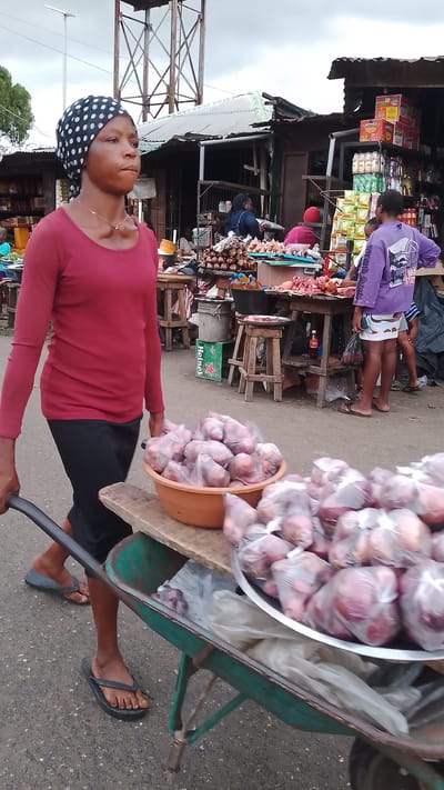 The image shows a street scene in Agbarho, Nigeria, likely a market. A woman wearing a red long-sleeved shirt and a black headscarf with white polka dots is pushing a wheelbarrow filled with produce. The wheelbarrow contains several plastic bags of red onions, some in a large orange bowl and others on a metal tray. In the background, there are market stalls with various goods, including food items like meat and bottled drinks. Several other people are visible in the background, engaged in market activities. The environment appears to be outdoors with a cloudy sky. The ground is paved.
