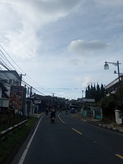 A road curves through a town under a cloudy sky. Several people on motorbikes travel along the asphalt road marked with yellow lines. Buildings and vegetation line both sides of the street, with a large advertisement on the left. Utility poles and wires crisscross overhead, adding to the urban landscape. A decorative street lamp stands on the right side of the road, further defining the scene.