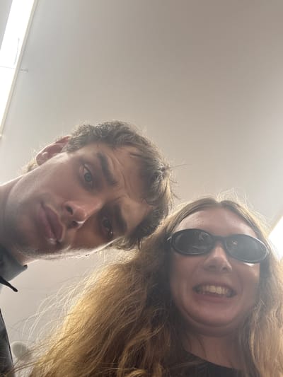 An informal, low-angle photograph features two individuals positioned above the camera. The male subject, on the left, has dark, wavy hair and light eyes. He exhibits a neutral expression with a slight furrow to his brow and his lips slightly parted. A portion of a dark garment is visible on his left shoulder. The female subject, on the right, possesses long, light brown, wavy hair and wears dark, oval sunglasses. She presents an exaggerated, wide-mouthed grin, revealing her teeth, with a visible double chin due to the upward camera angle. The background shows a light-colored ceiling illuminated by overhead fluorescent light fixtures. A thin wire or string is observable in the upper-left corner of the ceiling.